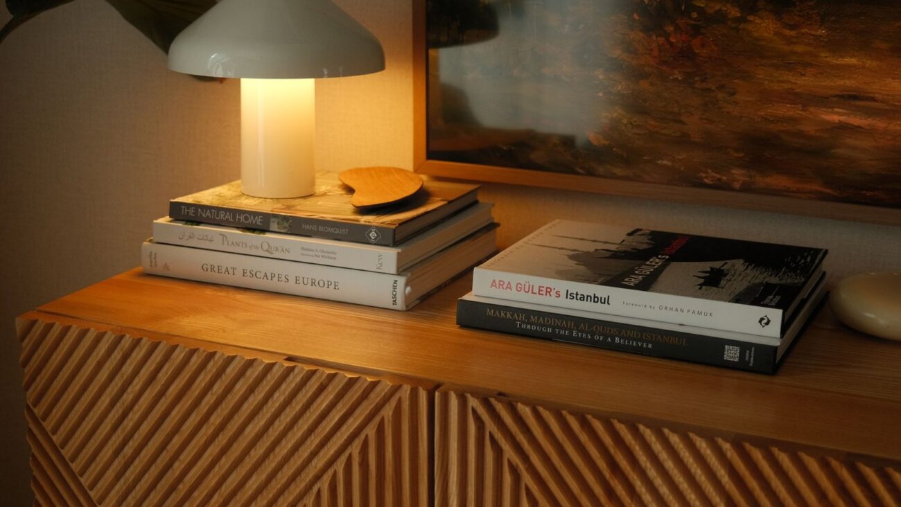 Mesa lateral de madeira com luminaria acesa e pilhas de livros em composicao acolhedora